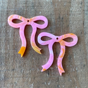 May include: Two flat bow-shaped charms featuring a marbled pattern of light pink, white, and golden-orange swirls. Each bow has a small circular hole at the top center for attachment, with smooth edges and a glossy finish on the front surface.