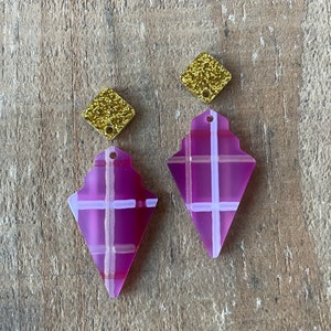 May include: A pair of purple and white plaid earrings with gold glitter accents. The earrings are shaped like a teardrop with a pointed bottom.
