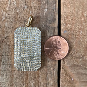 Puede incluir: Un colgante de oro con forma rectangular, completamente cubierto de pequeñas piedras transparentes. El colgante está al lado de un centavo estadounidense para comparar el tamaño.