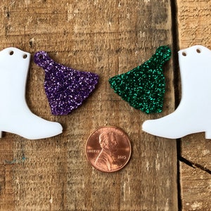 Puede incluir: Dos encantos blancos en forma de bota con un agujero en la parte superior. Un encanto está al lado de un encanto de purpurina morado y el otro al lado de un encanto de purpurina verde. Una moneda de un centavo está en el centro de la imagen para comparar el tamaño.