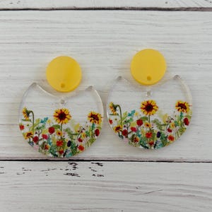 May include: A pair of dangle earrings featuring a clear, semi-circular acrylic base with a colourful embedded floral print including sunflowers, red poppies, and green foliage, topped with a solid, opaque yellow circular stud component.