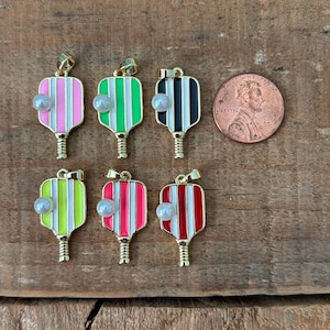May include: Six gold-toned pickleball paddle charms with colorful striped designs in pink, green, black, and red, each adorned with a faux pearl. A penny is included for size reference. The charms are displayed on a wooden surface.