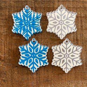 May include: Four snowflake-shaped ornaments arranged in a square pattern. Two ornaments are bright blue with a clear acrylic border, and two are white with a clear acrylic border. Each snowflake features a central circular detail and six points.