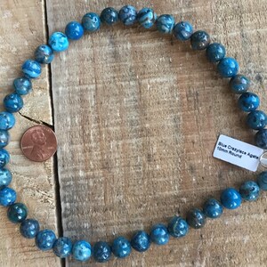 May include: A strand of blue crazy lace agate beads, 10mm round, on a wooden surface. A penny is included for size reference.