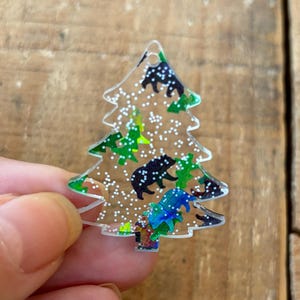 May include: A clear plastic Christmas tree ornament with a white glitter background and a green and black bear print.