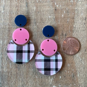 Puede incluir: Un par de pendientes de cuadros rosa y negro con detalles en rosa y azul marino. Los pendientes están hechos de acrílico y tienen una forma semicircular.