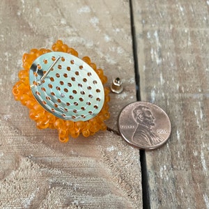 May include: Back view of an orange beaded brooch with a metal backing and a pin fastening. A UK penny is included for size reference.