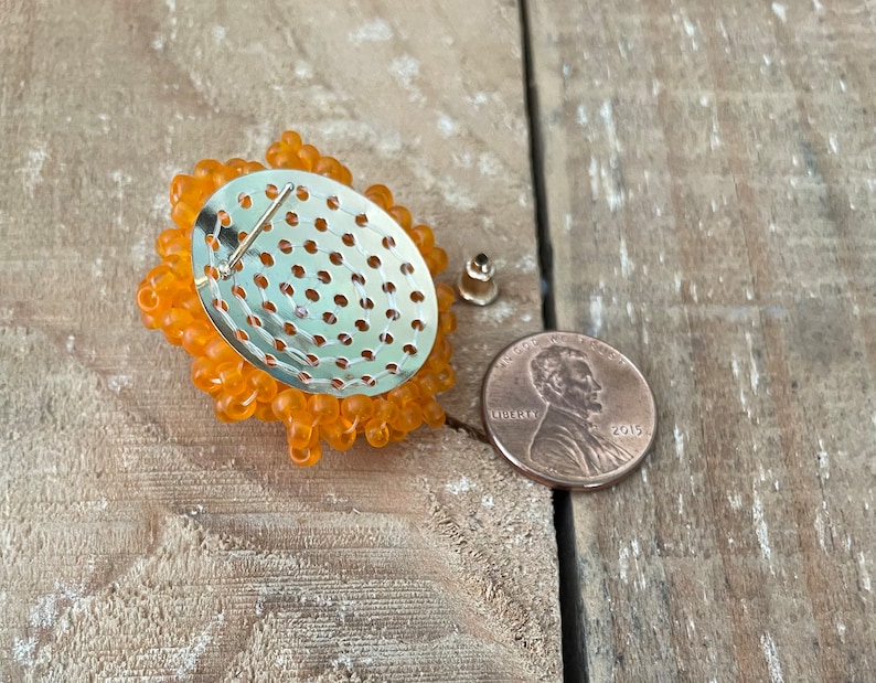 May include: Back view of an orange beaded brooch with a metal backing and a pin fastening. A UK penny is included for size reference.