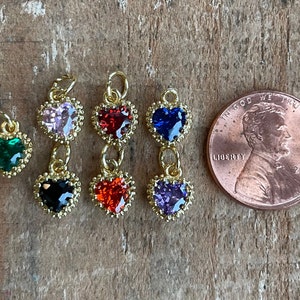 May include: Six gold heart-shaped charms with different colored gemstones. The charms are arranged in a row on a wooden surface. A US penny is included for scale.
