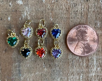 May include: Six gold heart-shaped charms with different colored gemstones. The charms are arranged in a row on a wooden surface. A US penny is included for scale.