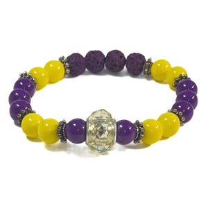 May include: A purple and yellow beaded bracelet with a clear crystal charm. The bracelet is made with lava rock beads and has silver accents.