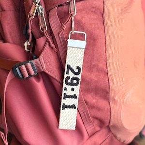 Puede incluir: Una mochila de color óxido con un llavero de tela blanca sujeto a un clip de metal plateado. El llavero presenta el texto bordado en negro 29:11 y está asegurado con un cierre rectangular de metal plateado, colgado de la cremallera.