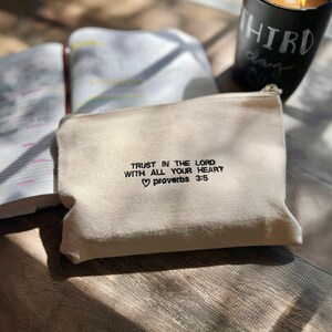 May include: A white canvas zippered pouch with the text "TRUST IN THE LORD WITH ALL YOUR HEART &hearts; Proverbs 3:5" embroidered in black.