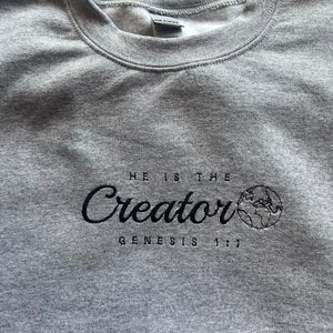 May include: Gray sweatshirt with black embroidered text that reads "He is the Creator Genesis 1:1". The text is in a cursive font and is accompanied by a small embroidered image of the Earth.