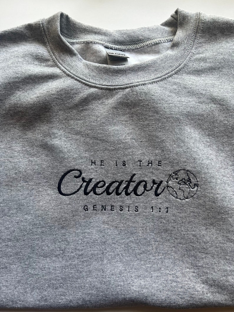 May include: Gray sweatshirt with black embroidered text that reads "He is the Creator Genesis 1:1". The text is in a cursive font and is accompanied by a small embroidered image of the Earth.