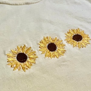 Puede incluir: Una camiseta blanca con tres girasoles bordados. Los girasoles son amarillos con centros marrones.