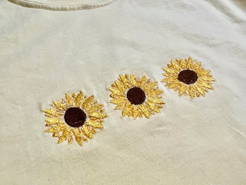 Puede incluir: Una camiseta blanca con tres girasoles bordados. Los girasoles son amarillos con centros marrones.
