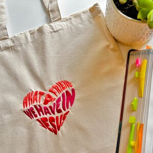 Op de afbeelding: Een witte canvas tote bag met een roze en rood geborduurd hart waarop staat "Wat een vriend hebben we in Jezus".