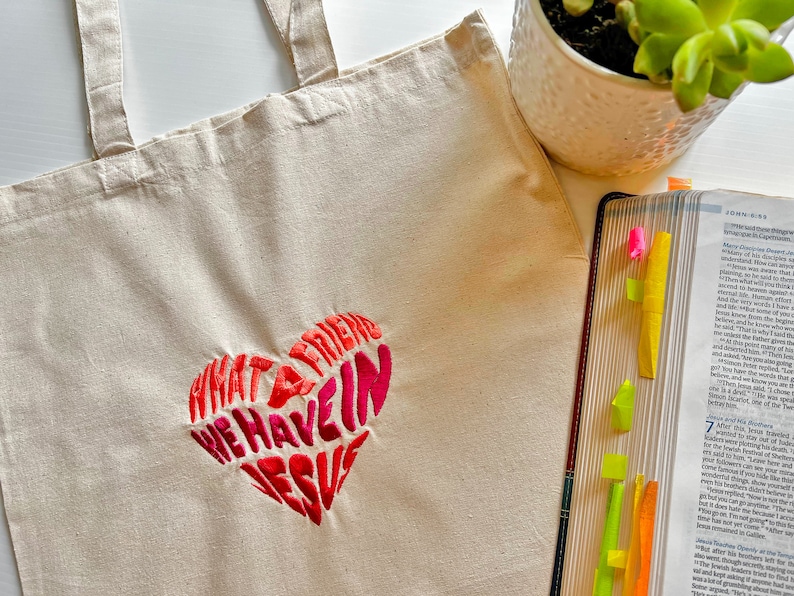 Op de afbeelding: Een witte canvas tote bag met een roze en rood geborduurd hart waarop staat "Wat een vriend hebben we in Jezus".