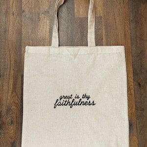 Puede incluir: Un bolso tote de lona beige con asas largas. Las palabras "great is thy faithfulness" est&aacute;n impresas en escritura negra en la parte delantera. La bolsa est&aacute; hecha de una tela resistente y es adecuada para transportar varios art&iacute;culos.
