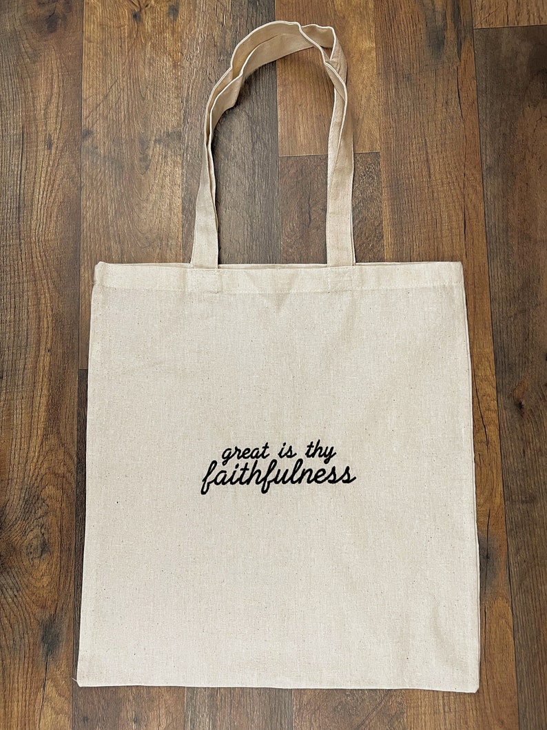 Puede incluir: Un bolso tote de lona beige con asas largas. Las palabras "great is thy faithfulness" est&aacute;n impresas en escritura negra en la parte delantera. La bolsa est&aacute; hecha de una tela resistente y es adecuada para transportar varios art&iacute;culos.