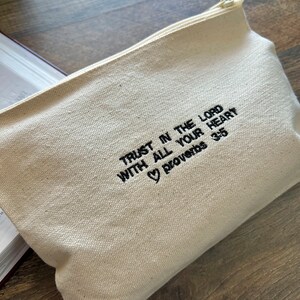 Peut inclure: Une pochette zipp&eacute;e en toile blanche avec une broderie noire qui dit "Trust in the Lord with all your heart Proverbs 3:5" avec un symbole de c&oelig;ur avant le mot "Proverbs".