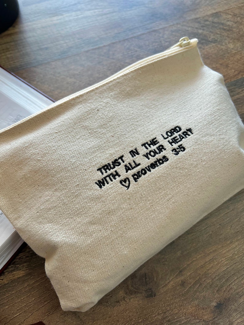 Peut inclure: Une pochette zipp&eacute;e en toile blanche avec une broderie noire qui dit "Trust in the Lord with all your heart Proverbs 3:5" avec un symbole de c&oelig;ur avant le mot "Proverbs".