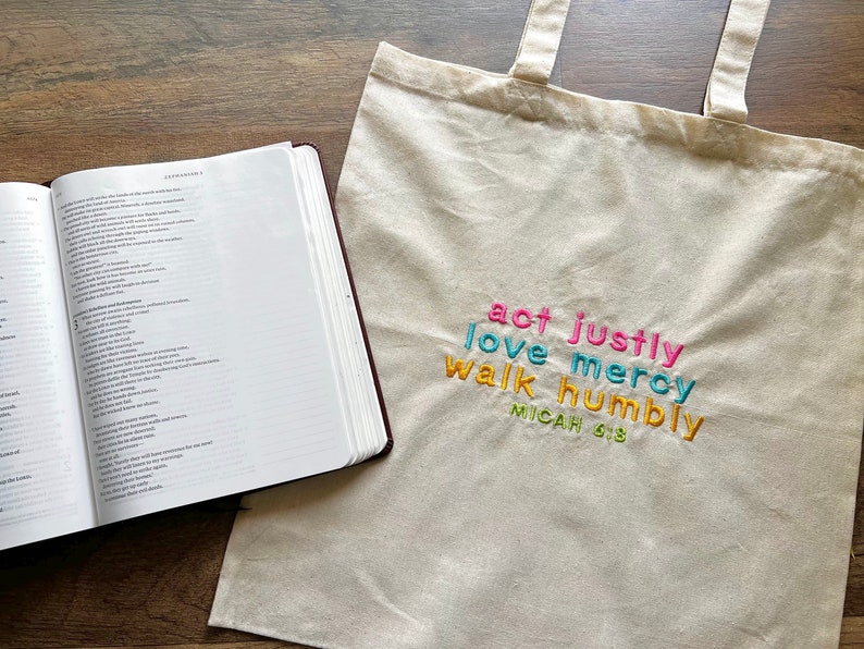 May include: A white canvas tote bag with the words "act justly, love mercy, walk humbly" embroidered in colorful letters. The text "Micah 6:8" is embroidered in yellow below the other words.