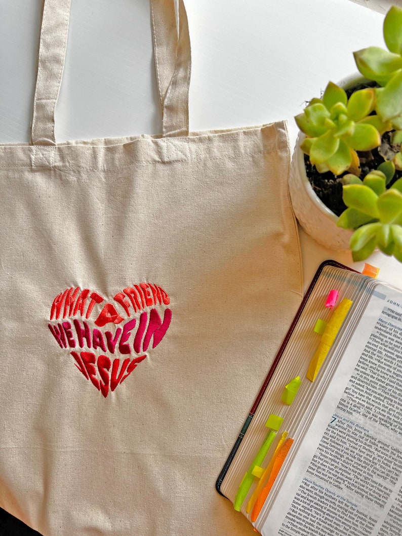 Op de afbeelding: Een witte canvas tote bag met een rood en roze geborduurd hart waarop staat "Wat een vriend hebben we in Jezus".