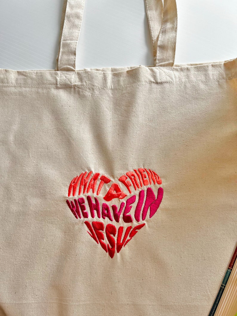 Op de afbeelding: Een witte canvas tote bag met een rood geborduurd hart waarop staat "Wat een vriend hebben we in Jezus".