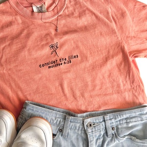 May include: A peach-colored t-shirt with a floral design and the text "consider the lilies matthew 6:28" embroidered on the front.