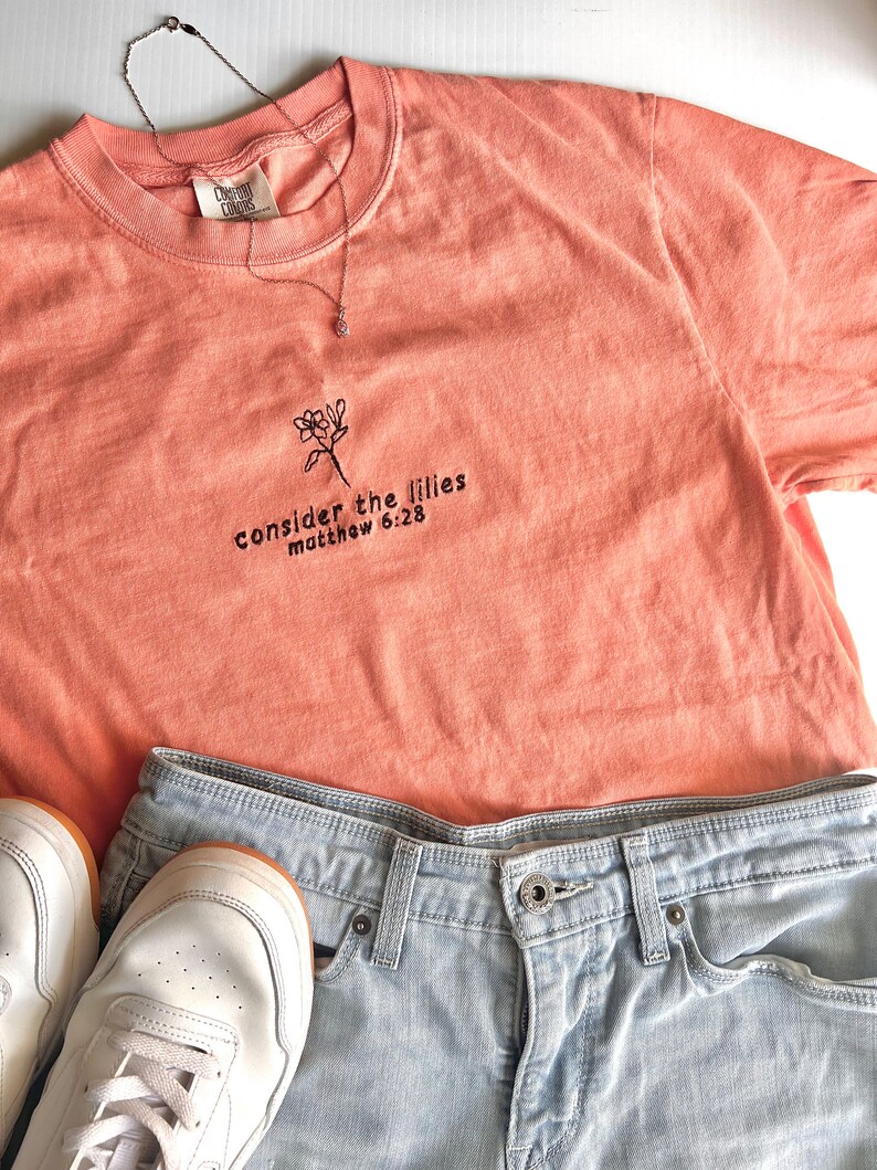 May include: A peach-colored t-shirt with a floral design and the text "consider the lilies matthew 6:28" embroidered on the front.