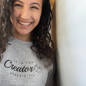 May include: A woman with curly brown hair is smiling and wearing a gray sweatshirt with the text "He is the Creator Genesis 1:1" and a small graphic of the Earth.
