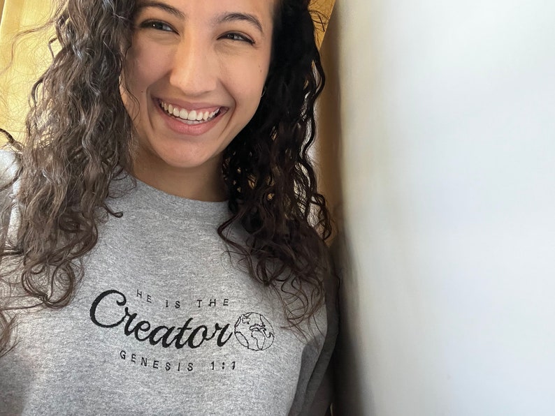 May include: A woman with curly brown hair is smiling and wearing a gray sweatshirt with the text "He is the Creator Genesis 1:1" and a small graphic of the Earth.