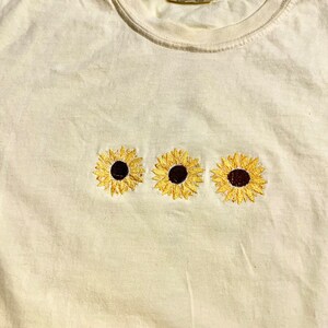 Puede incluir: Una camiseta de color amarillo claro con tres girasoles bordados. Los girasoles son amarillos con centros marrones.