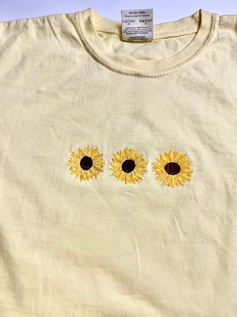Puede incluir: Una camiseta de color amarillo claro con tres girasoles bordados. Los girasoles son amarillos con centros marrones.
