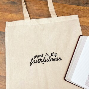 Puede incluir: Bolso tote de color crema con el texto bordado "great is thy faithfulness" en escritura negra. El bolso tiene asas de color crema a juego. Un libro encuadernado en cuero marr&oacute;n est&aacute; al lado del bolso.