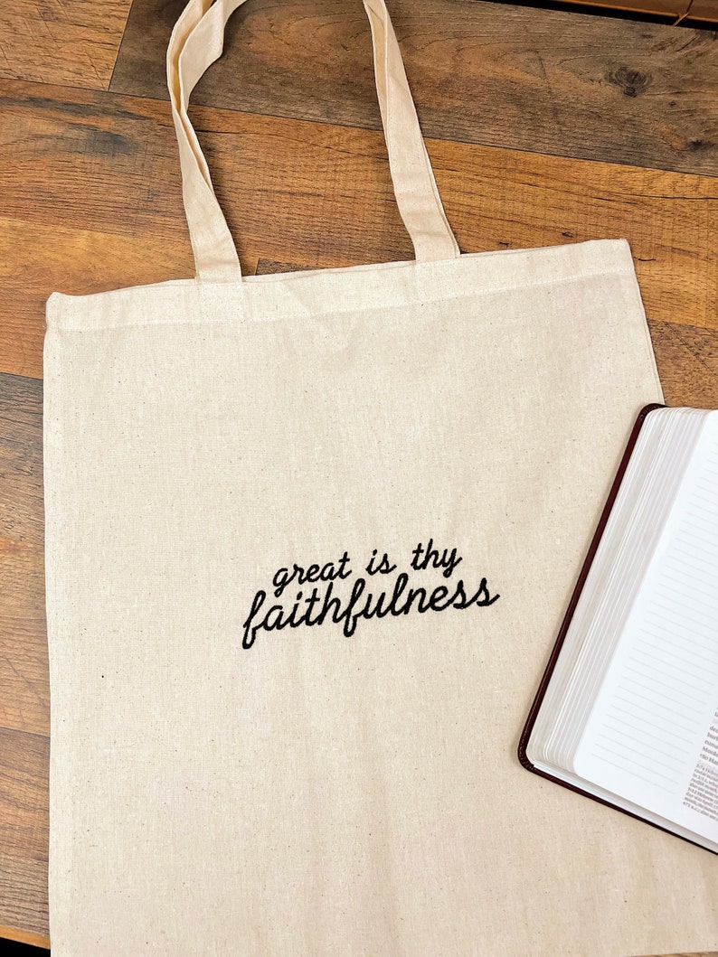Puede incluir: Bolso tote de color crema con el texto bordado "great is thy faithfulness" en escritura negra. El bolso tiene asas de color crema a juego. Un libro encuadernado en cuero marr&oacute;n est&aacute; al lado del bolso.