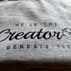 May include: Gray sweatshirt with black embroidered text that reads "He is the Creator Genesis 1:1" with a small embroidered globe to the right of the text.