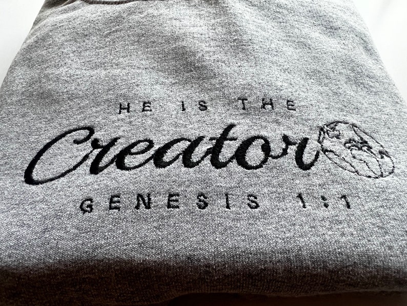 May include: Gray sweatshirt with black embroidered text that reads "He is the Creator Genesis 1:1" with a small embroidered globe to the right of the text.