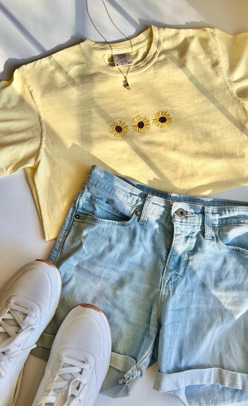 Puede incluir: Una camiseta amarilla con tres girasoles bordados en la parte delantera. La camiseta se combina con pantalones cortos de mezclilla azul claro y zapatillas blancas.