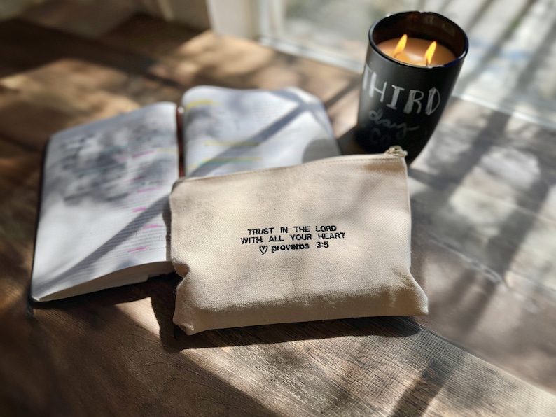 May include: A white canvas zippered pouch with the text "TRUST IN THE LORD WITH ALL YOUR HEART &hearts; Proverbs 3:5" embroidered in black.