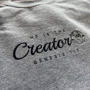 May include: A gray sweatshirt with black embroidered text that reads "He is the Creator Genesis 1:1" with a small embroidered globe to the right of the text.