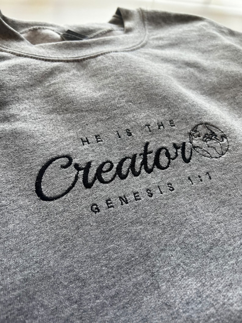 May include: A gray sweatshirt with black embroidered text that reads "He is the Creator Genesis 1:1" with a small embroidered globe to the right of the text.