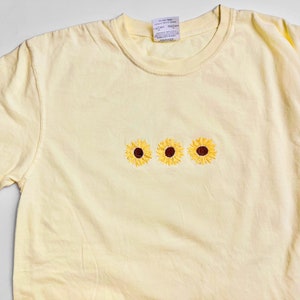 Puede incluir: Una camiseta de color amarillo p&aacute;lido con tres girasoles bordados en fila.