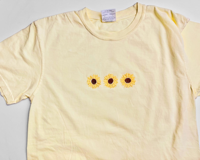 Puede incluir: Una camiseta de color amarillo p&aacute;lido con tres girasoles bordados en fila.