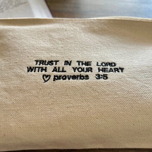 Peut inclure: Une pochette zipp&eacute;e en toile blanche avec un texte brod&eacute; en noir qui dit "TRUST IN THE LORD WITH ALL YOUR HEART &hearts; Proverbs 3:5".