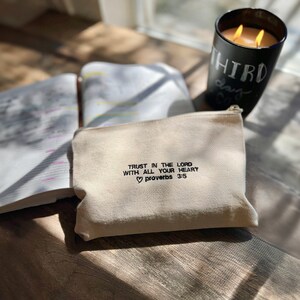 Peut inclure: Une pochette zipp&eacute;e en toile beige avec le texte "TRUST IN THE LORD WITH ALL YOUR HEART  proverbs 3:5" brod&eacute; en noir. La pochette est pos&eacute;e sur une surface en bois &agrave; c&ocirc;t&eacute; d'un livre ferm&eacute; et d'une bougie noire.