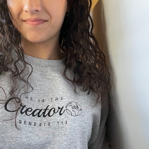 May include: Gray sweatshirt with the text "He is the Creator" and "Genesis 1:1" in black lettering. A small graphic of the Earth is next to the word "Creator".