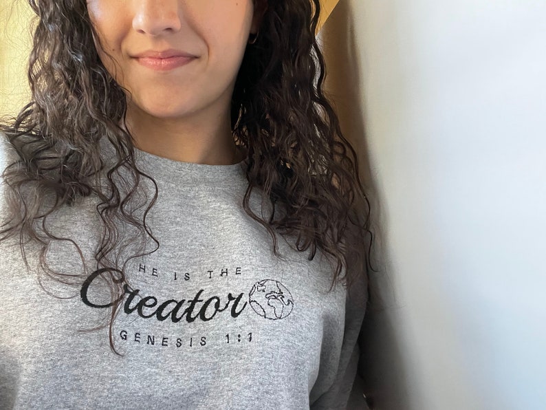 May include: Gray sweatshirt with the text "He is the Creator" and "Genesis 1:1" in black lettering. A small graphic of the Earth is next to the word "Creator".